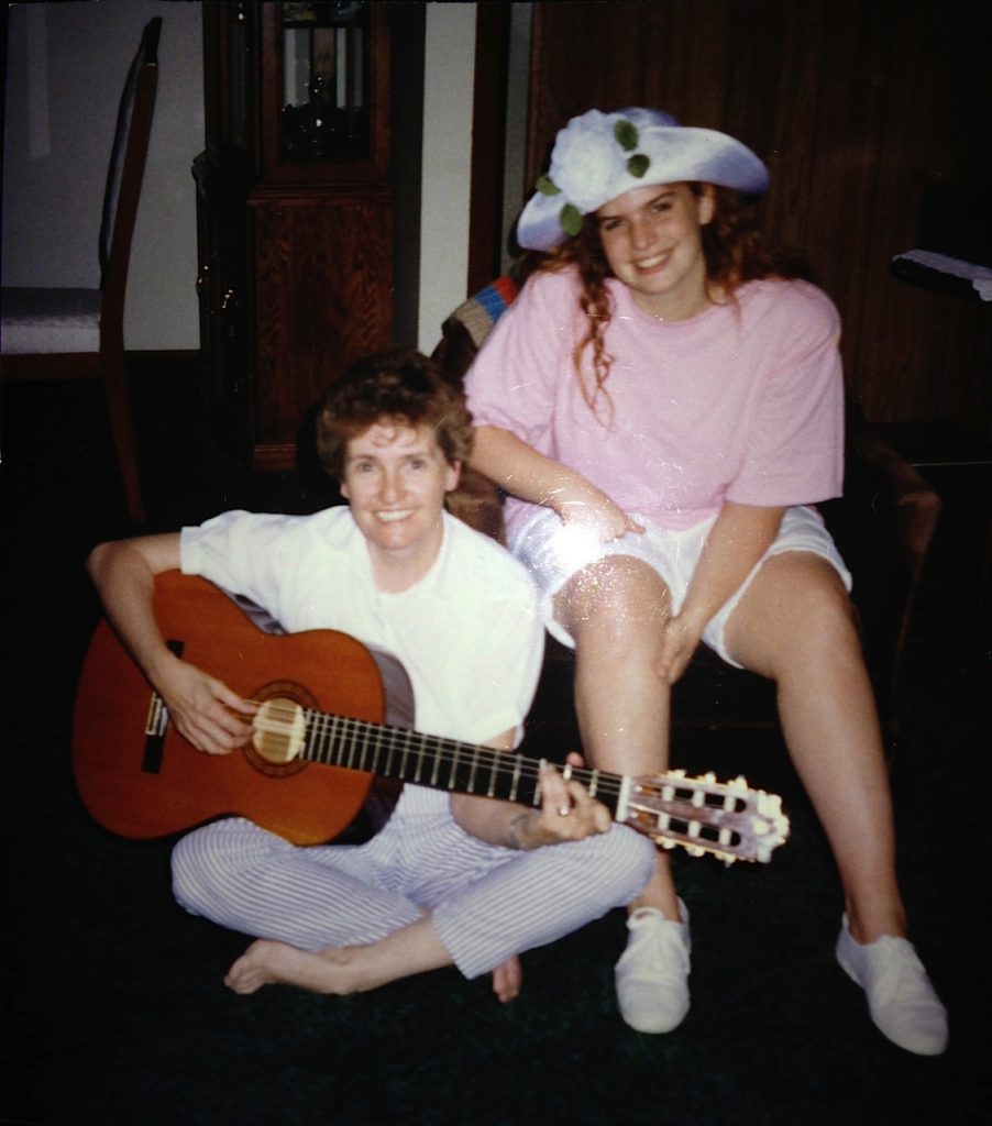 Gaile sitting on the floor holding her guitar with me sitting in a chair wearing in a large white hat with a flower, oversized pink t-shirt, white shorts and white sneakers
